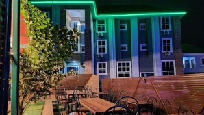An outdoor seating area with wooden tables and metal chairs, surrounded by a building with multiple windows and air conditioning units. - LAGENDA HOTELS (Enugu)