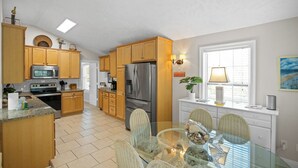 Una cocina con armarios de madera, electrodomésticos de acero inoxidable, una mesa de comedor de vidrio, sillas de mimbre y una ventana con persianas.