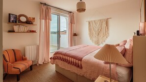 Ein Schlafzimmer mit Bett, Stuhl, Nachttisch, Lampe, Heizkörper, einem Regal mit Büchern und Dekoration sowie Blick durch das Fenster.