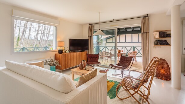 Ein modernes Wohnzimmer mit einer Couch, Sesseln, einem Fernseher und einem Balkon mit Gartenmöbeln.