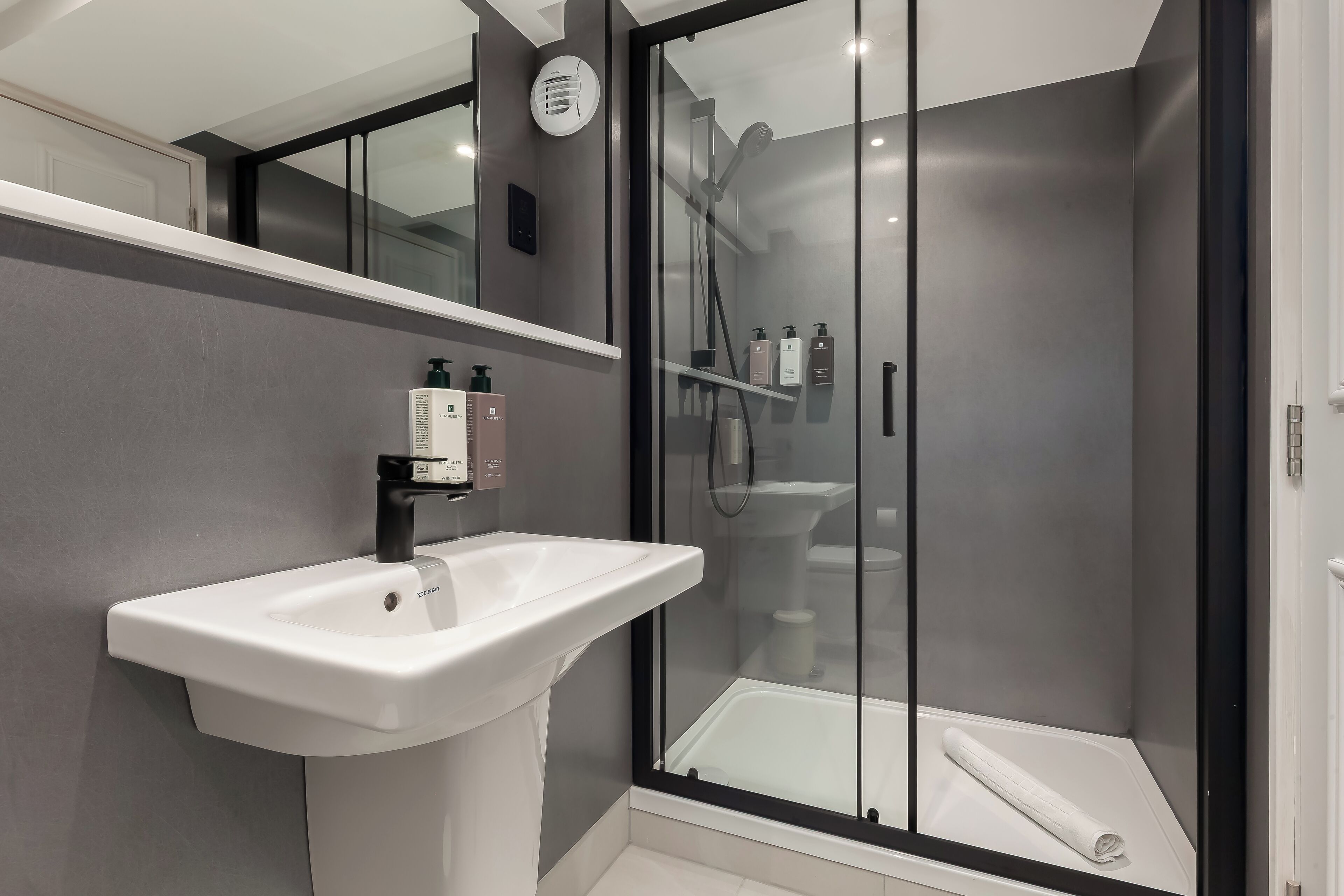 Un baño moderno con un lavamanos pedestal blanco, una ducha con cerramiento de vidrio y una toalla blanca en el suelo.