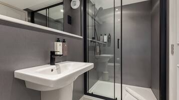 Un baño moderno con un lavamanos pedestal blanco, una ducha con cerramiento de vidrio y una toalla blanca en el suelo.