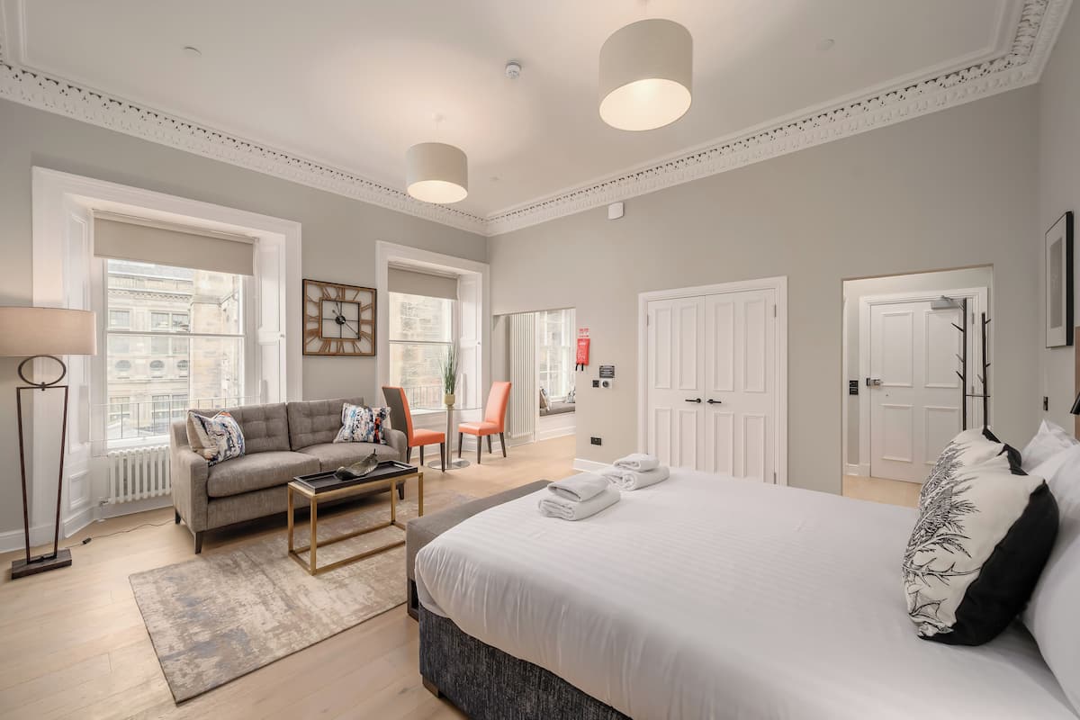 Une chambre d’hôtel moderne avec un grand lit, un canapé, une table basse et un bureau. Elle comprend deux portes, une fenêtre et un radiateur.