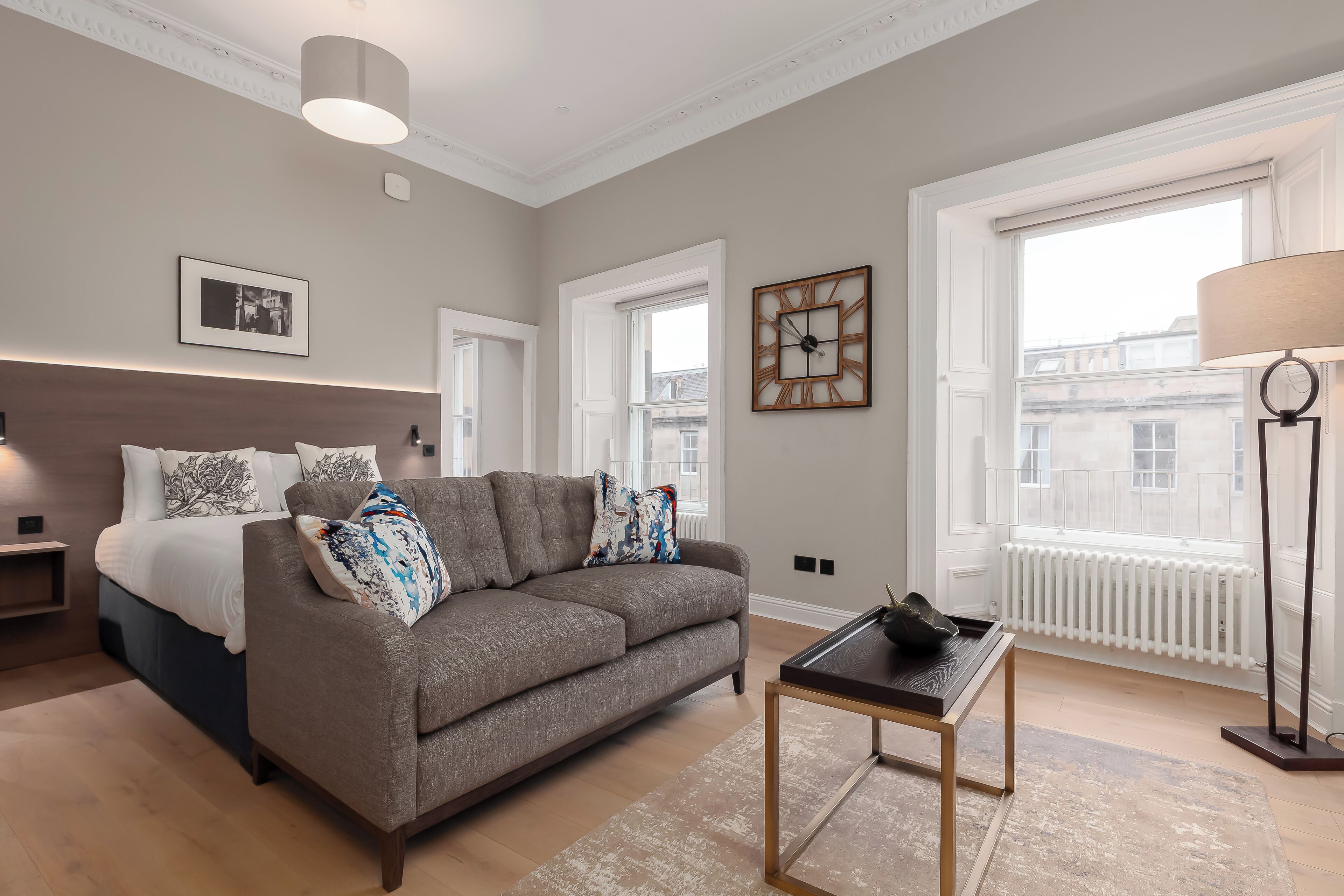 A modern bedroom with a grey sofa, a wooden nightstand, a bedside table, and a large window with a view of buildings.