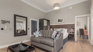 Een moderne woonkamer met een grijze bank, een salontafel en een grote spiegel. Achterin staat een bed met hoofdbord en een klein tafeltje met een stoel.