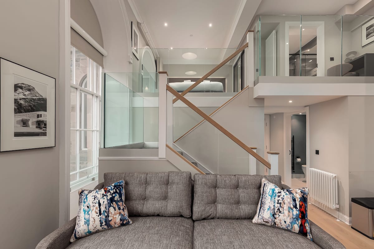 Un salon moderne avec un canapé gris, des coussins colorés, un escalier en verre et un tableau encadré au mur.