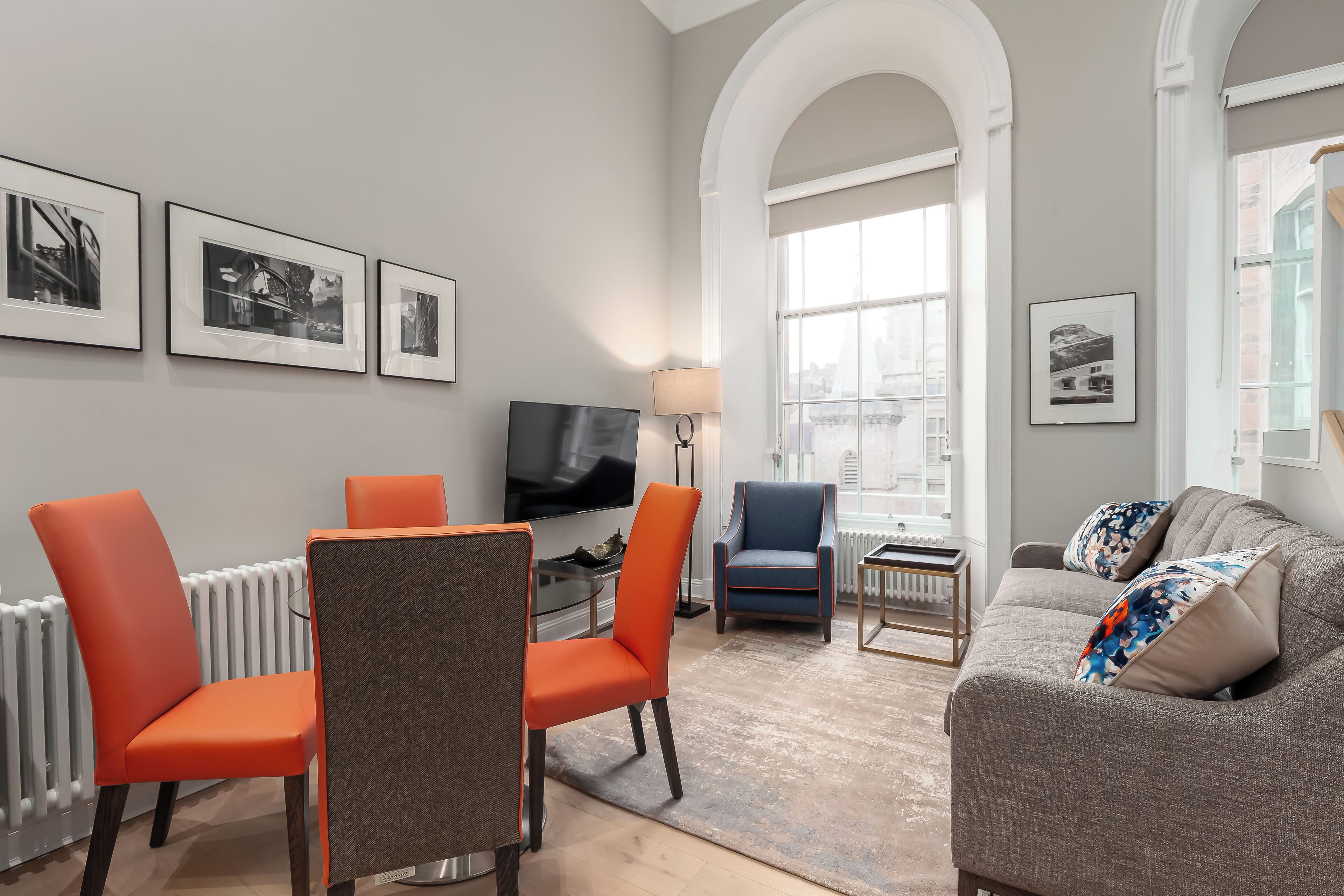 A modern living room with a grey sofa, orange and blue chairs, a flat-screen TV, and framed pictures on the wall.
