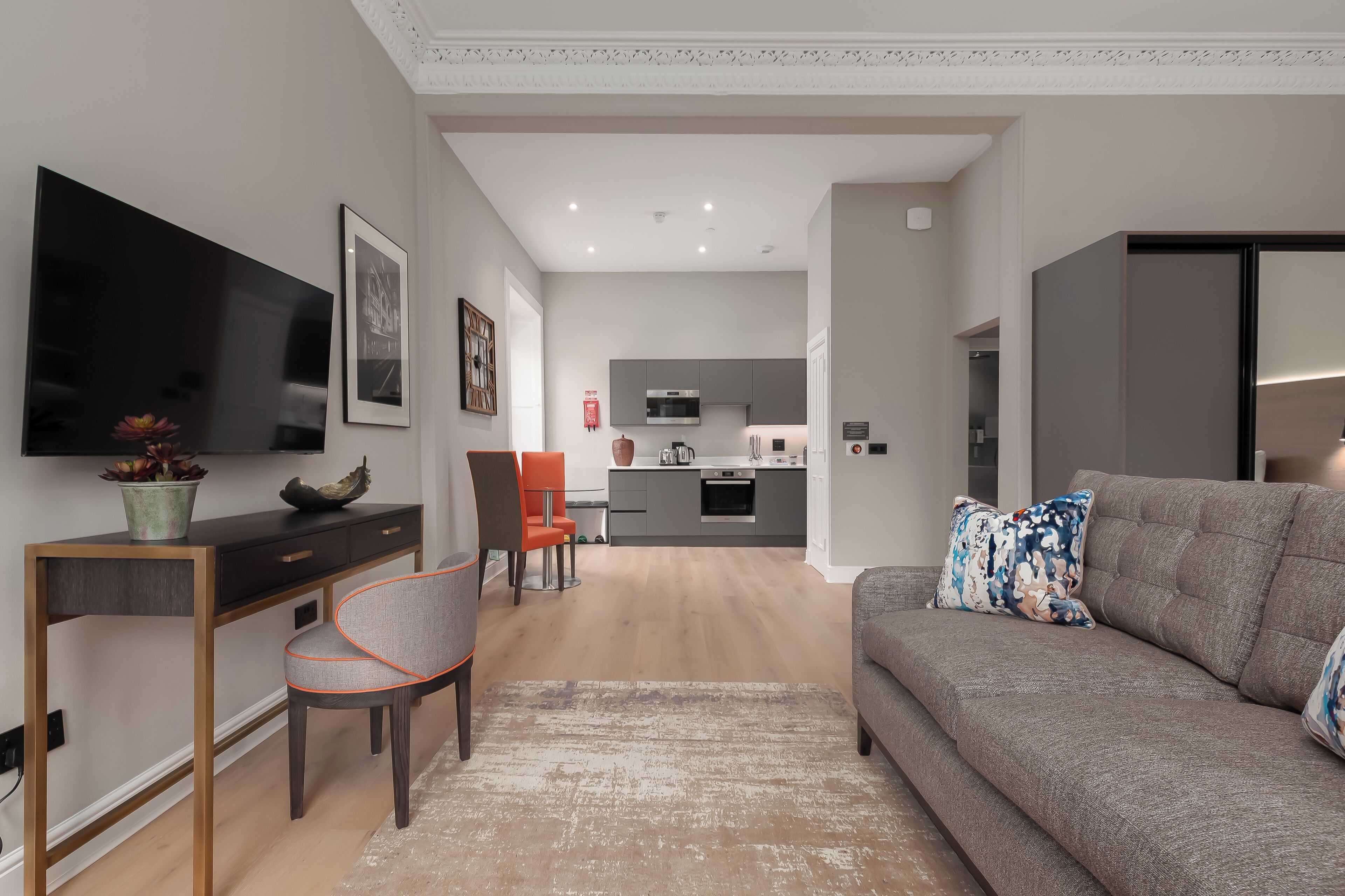 A modern living room with a grey sofa, a flat-screen TV, a dining area with a chair, and a kitchen in the background.