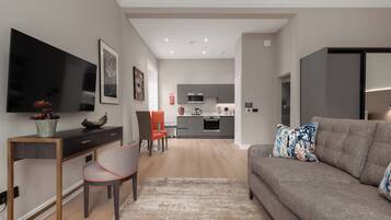 A modern living room with a grey sofa, a flat-screen TV, a dining area with a chair, and a kitchen in the background.
