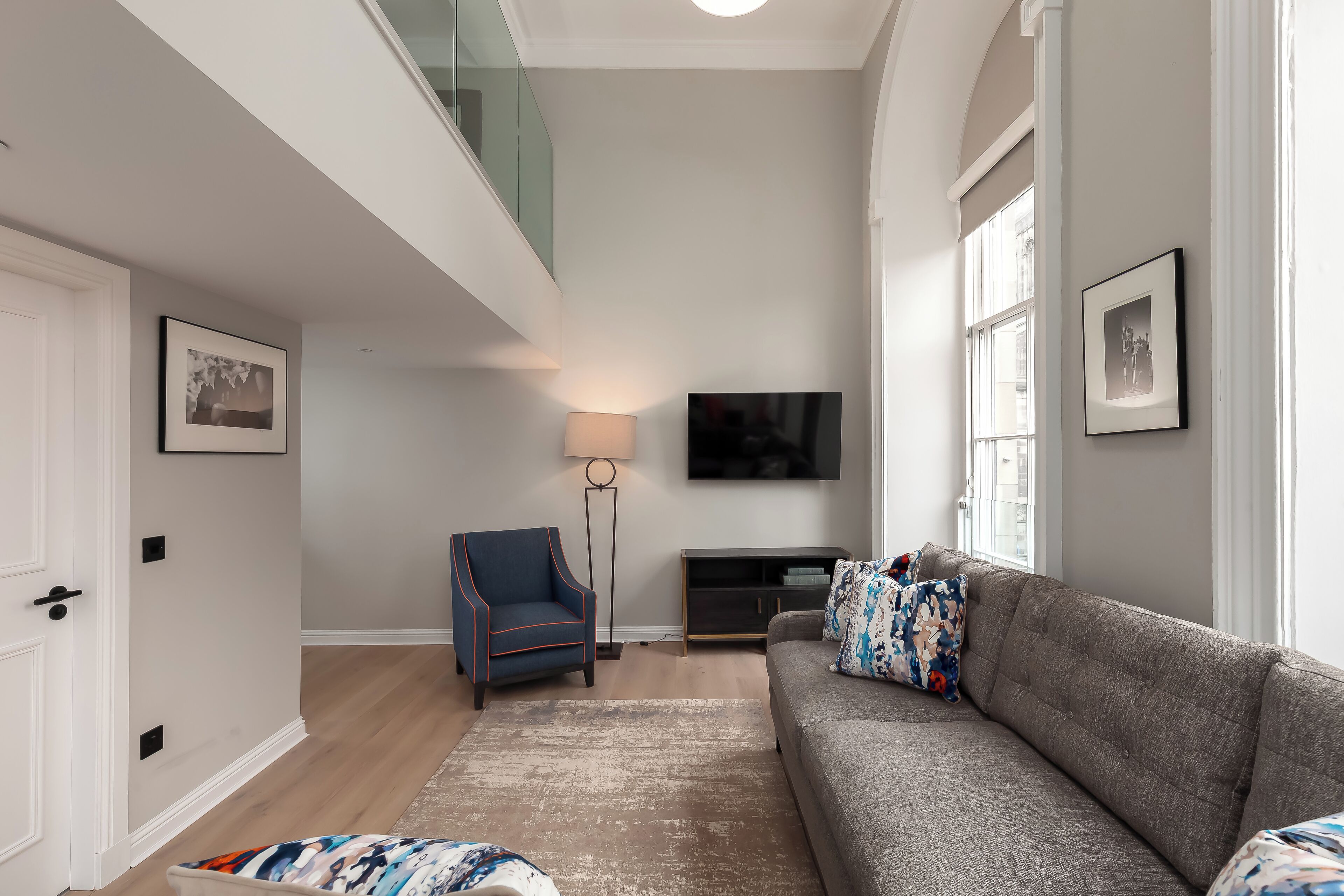 A modern living room with a grey sofa, a blue armchair, a floor lamp, and a flat-screen TV mounted on the wall.