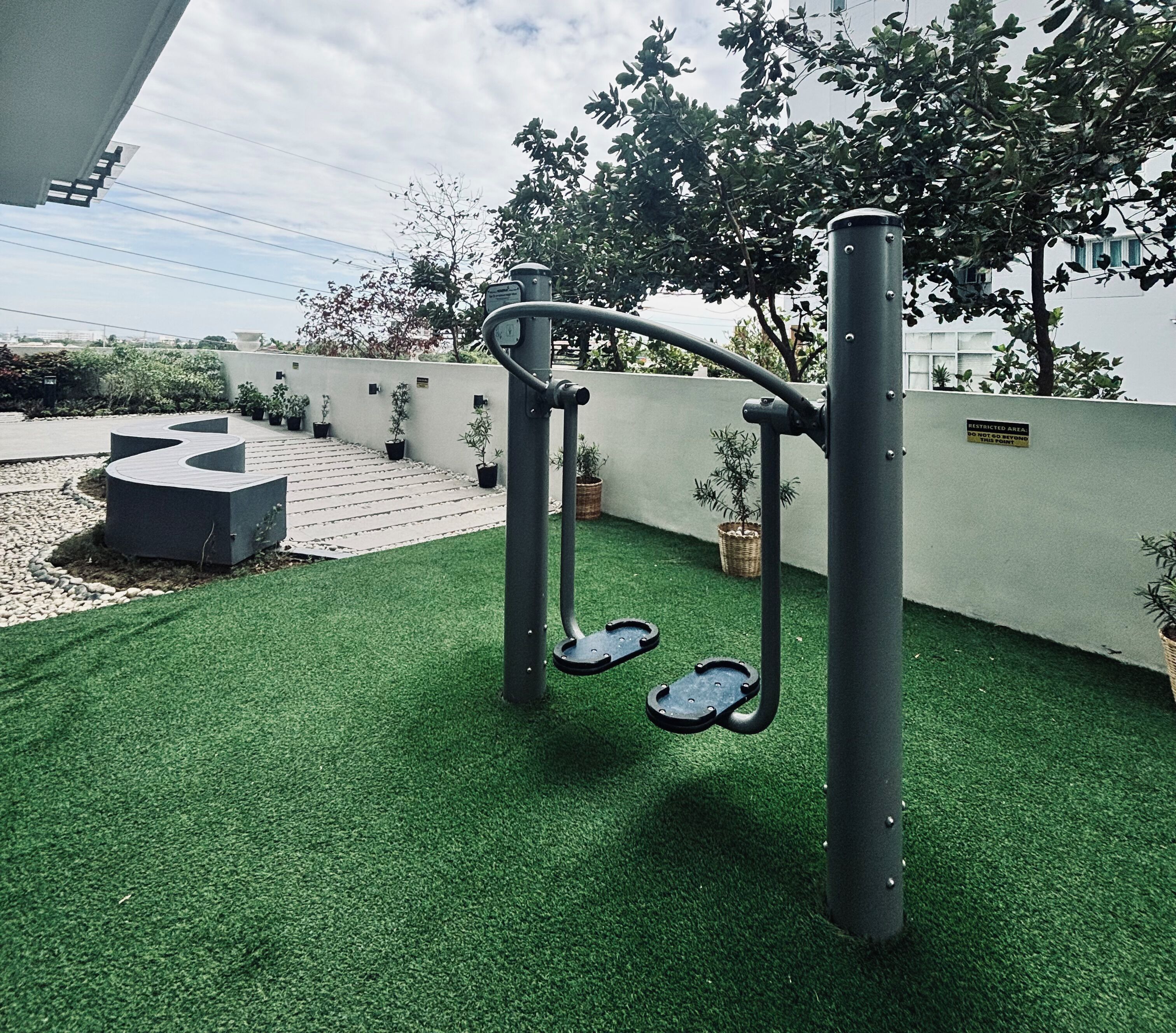Zona de ejercicios al aire libre con barra de dominadas y pedales para pies, rodeada de césped artificial verde y plantas en macetas.