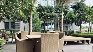 Un área de patio al aire libre con mobiliario de mimbre, incluyendo mesas y sillas, rodeado de vegetación y un edificio moderno al fondo.