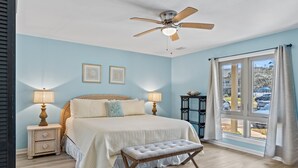 Un dormitorio con cama, lámparas de noche, ventilador de techo, ventana con cortinas y vistas al exterior.