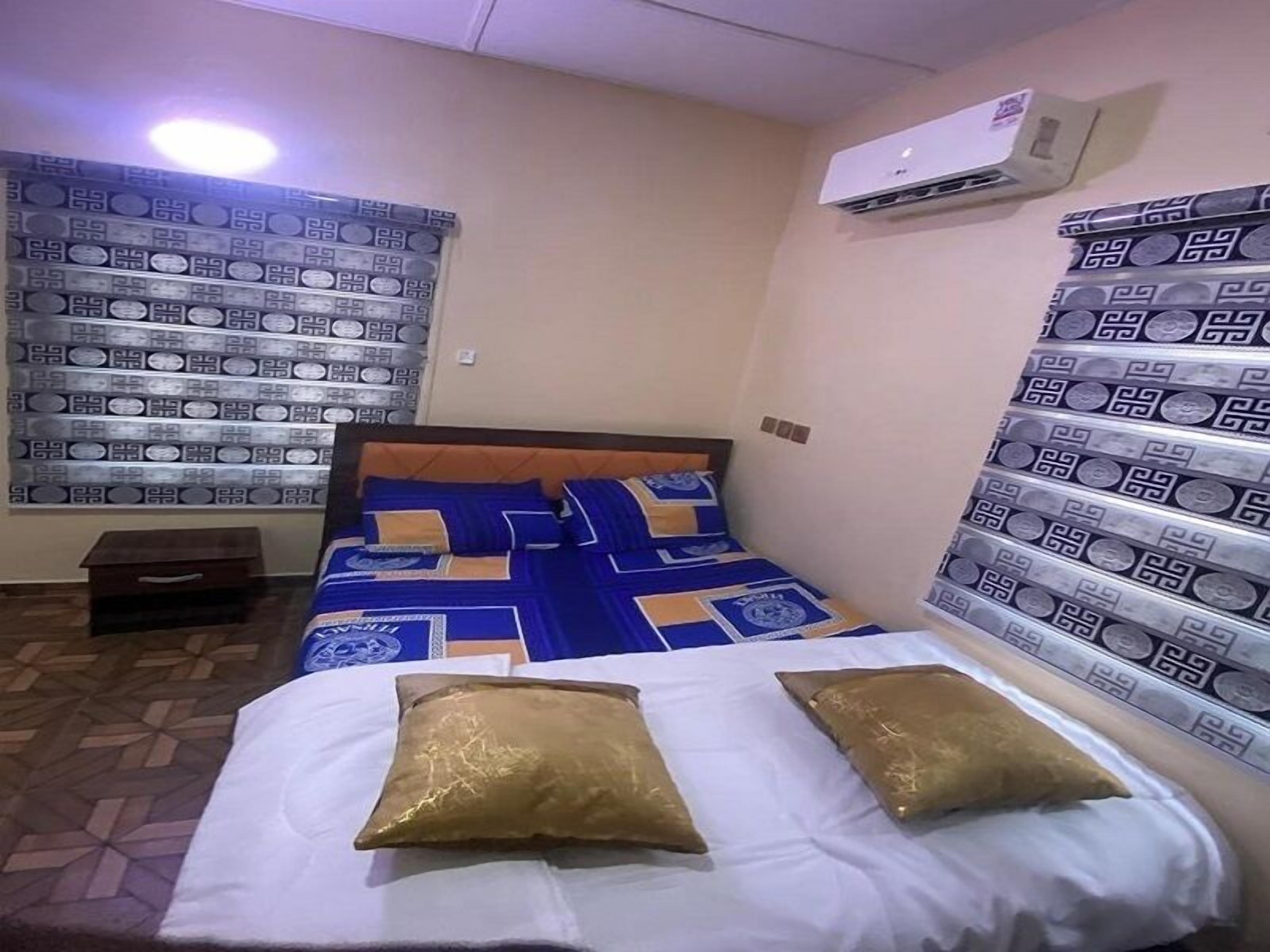 A hotel room with a bed, a nightstand, an air conditioning unit, patterned curtains, and a tiled floor.