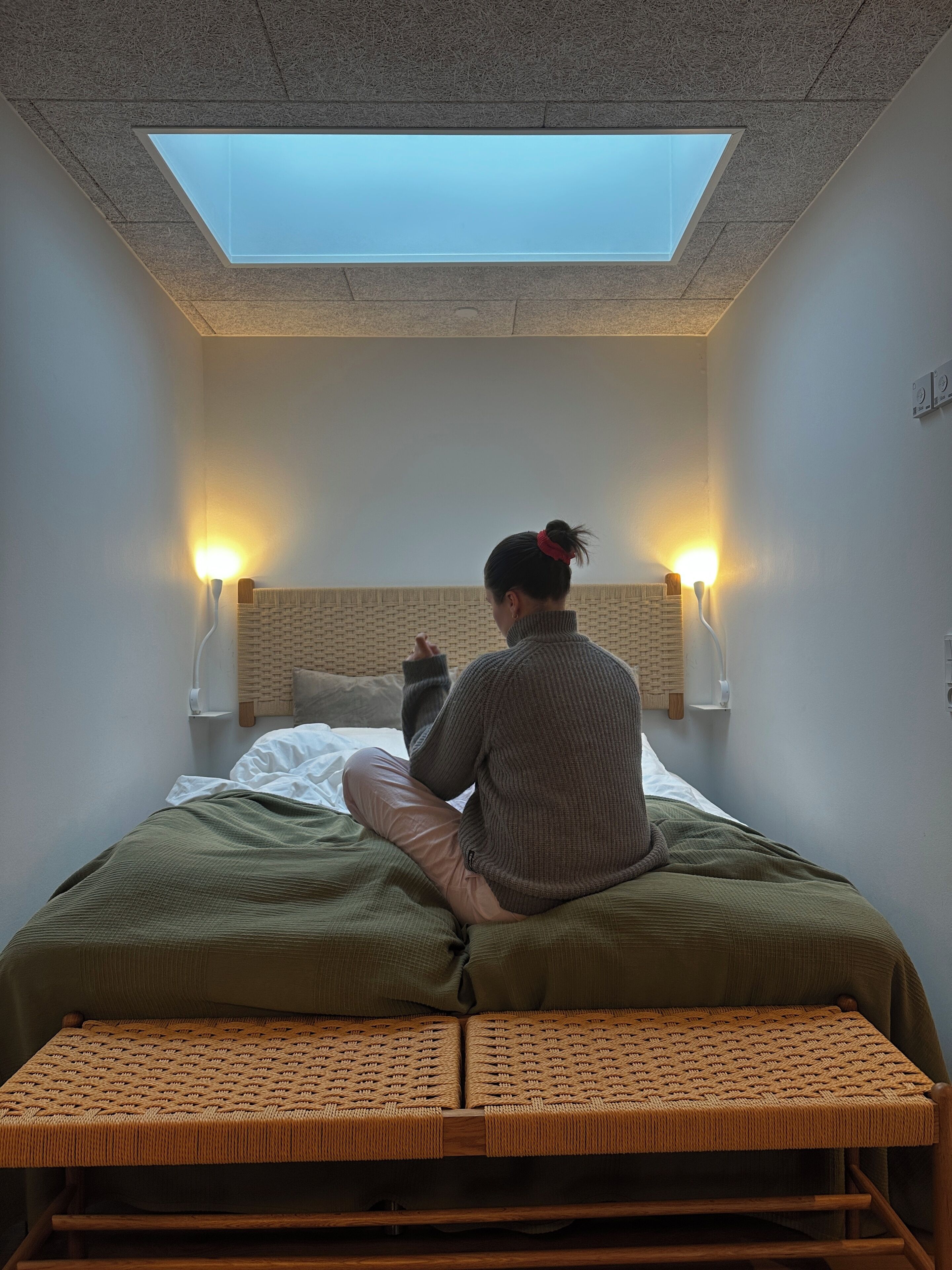 Ein Zimmer mit Oberlicht, einem Bett mit grüner Bettdecke, einer gewebten Bank und zwei Wandlampen.