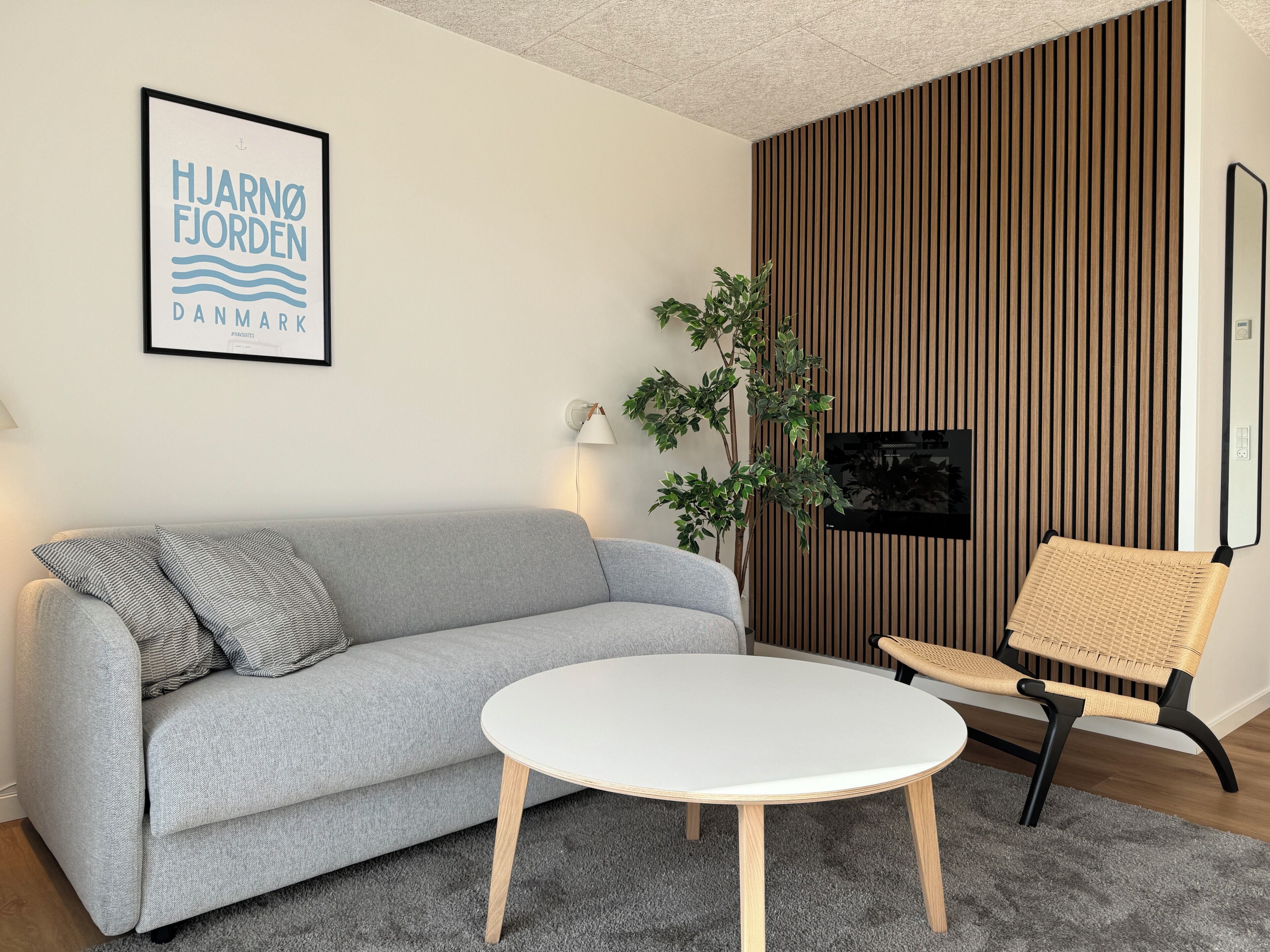 Een moderne woonkamer met een grijze bank, een ronde witte tafel, een rieten stoel, een aan de muur gemonteerde televisie en een lijst met een tekst.