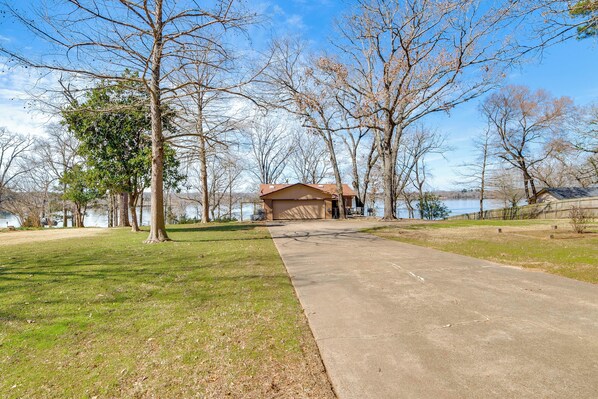 A concrete pathway leading to a building with a brown roof, surrounded by trees and a body of water. - Winnsboro Lake Retreat: On-site Swimming & Fishing (Winnsboro)