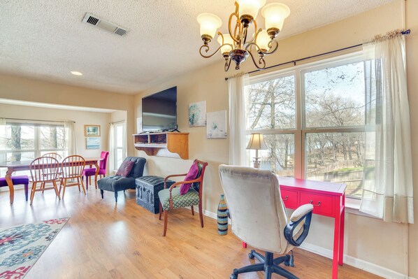 A spacious living room with a wooden floor, a red desk, a comfortable chair, a television, and a large window with a view of trees. - Winnsboro Lake Retreat: On-site Swimming & Fishing (Winnsboro)