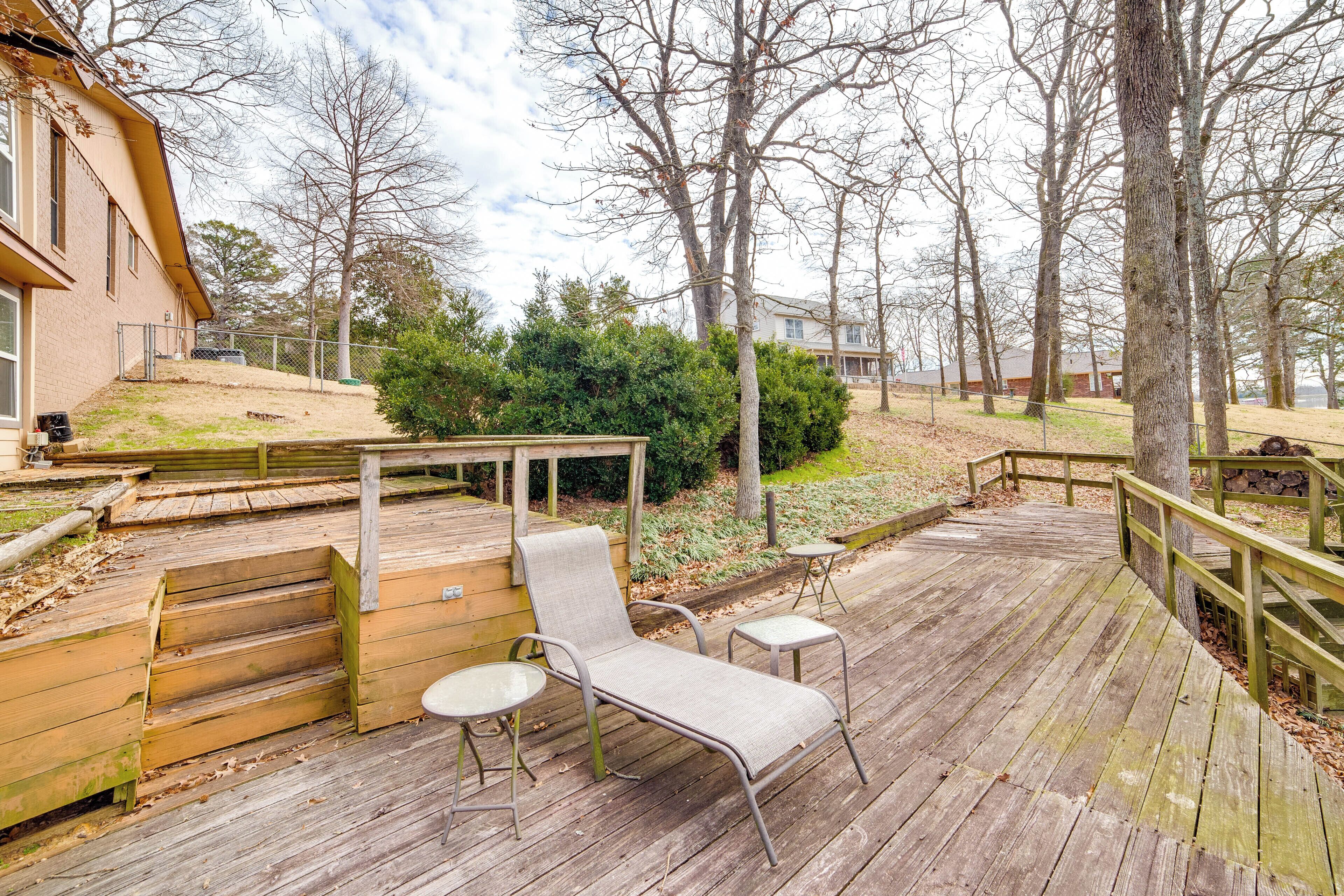 A wooden deck with a lounge chair and two small tables, surrounded by trees and a building.