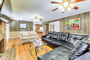 A living room with a black leather sofa, a floral armchair, and a glass coffee table. There is a ceiling fan and a painting on the wall.