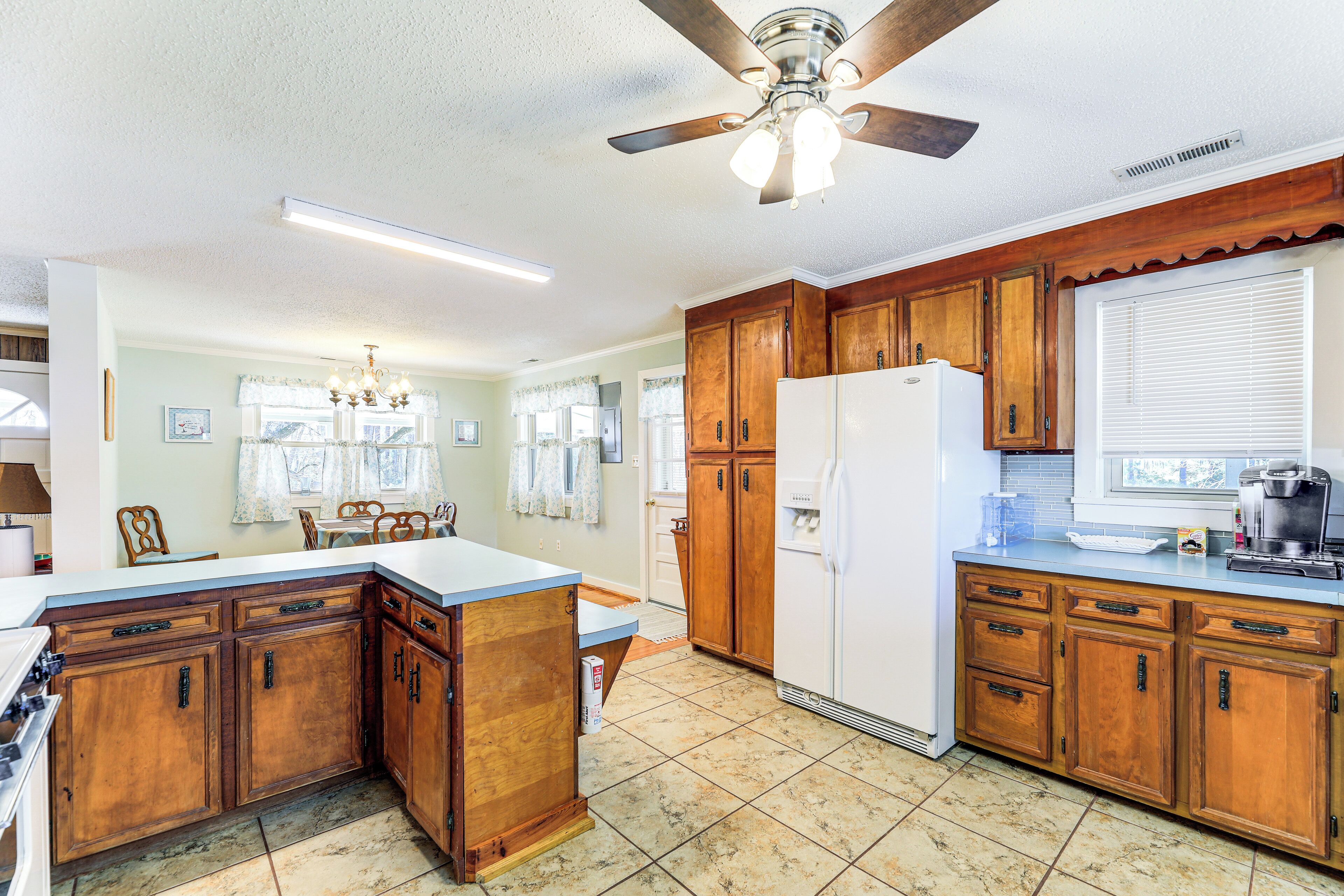 A kitchen with wooden cabinets, a white refrigerator, a ceiling fan, and a window with blinds.
