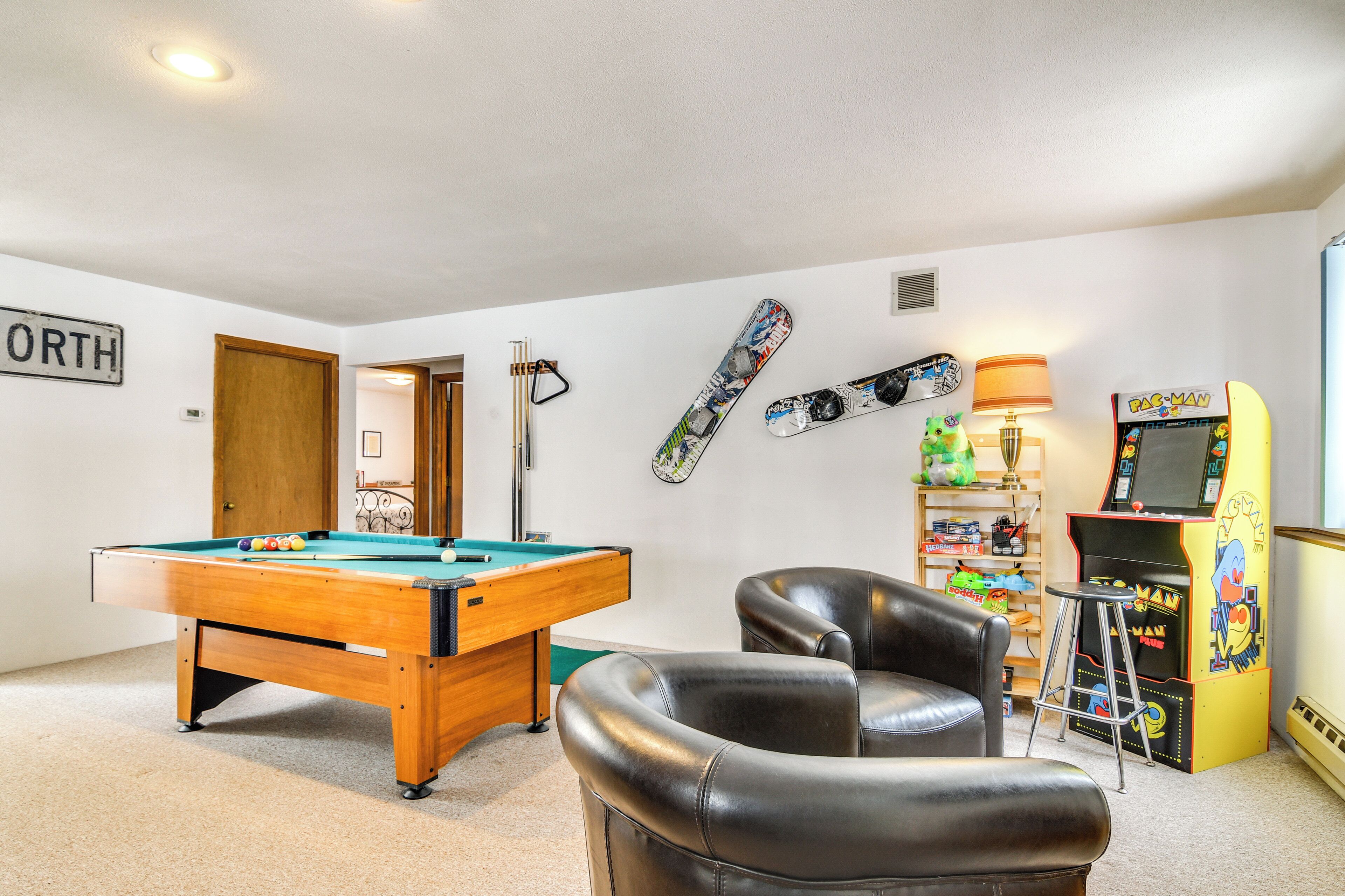 Een kamer met een pooltafel, twee leren fauteuils, een dartbord, een skateboard en een Pac-Man spelcomputer.