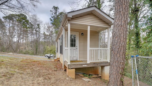 Een klein, laag huis met een veranda, omgeven door bomen en een gaashek.