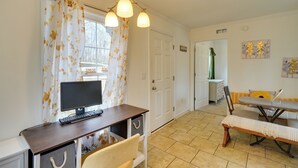 Een kamer met een bureau, stoel en een eettafel met banken. Er is een deur naar een andere kamer en een raam met gordijnen.
