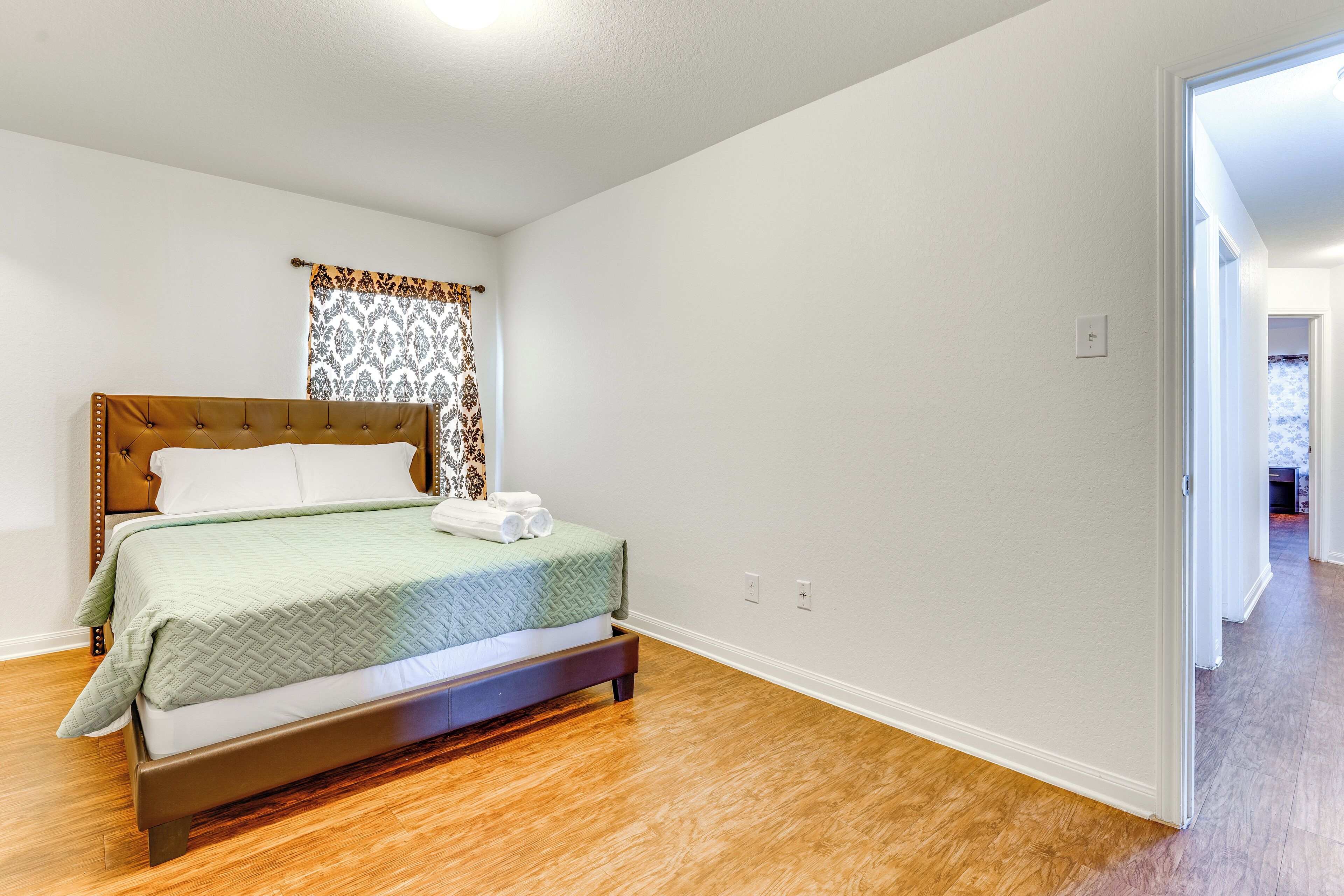 Un dormitorio con cama, suelo de madera, paredes blancas y cortina estampada.