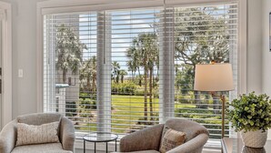 A room with two armchairs, a small table, a lamp, and a view of a green landscape through large windows with blinds. - Oceanfront beach villa in Palmetto Dunes (Hilton Head)