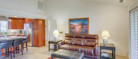 A modern living room with a leather sofa, a dining area with a kitchen in the background, and a painting on the wall.