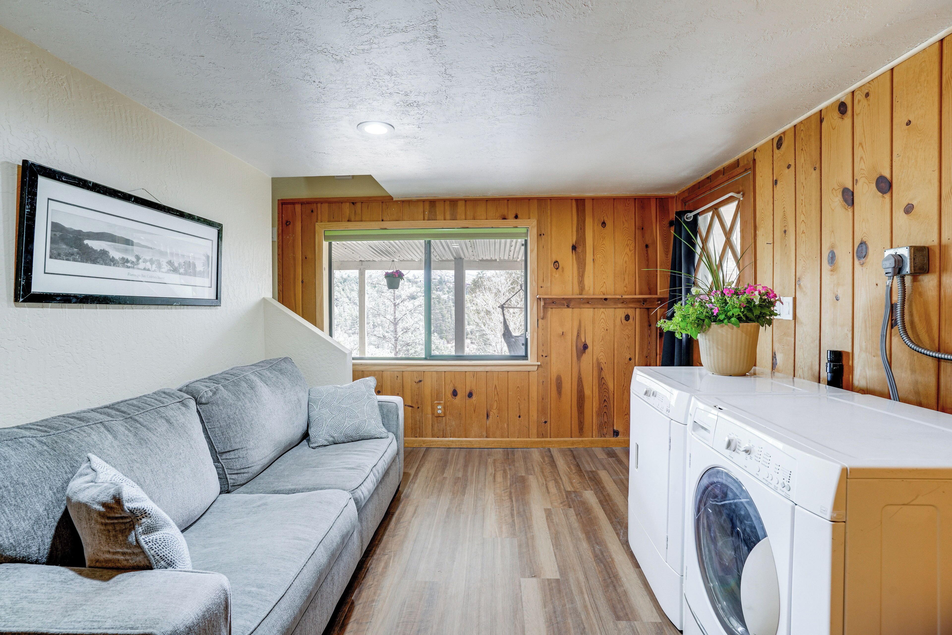 Ett kompakt bostadsutrymme med en grå soffa, träväggar, en tvättmaskin och ett fönster med utsikt över träd.