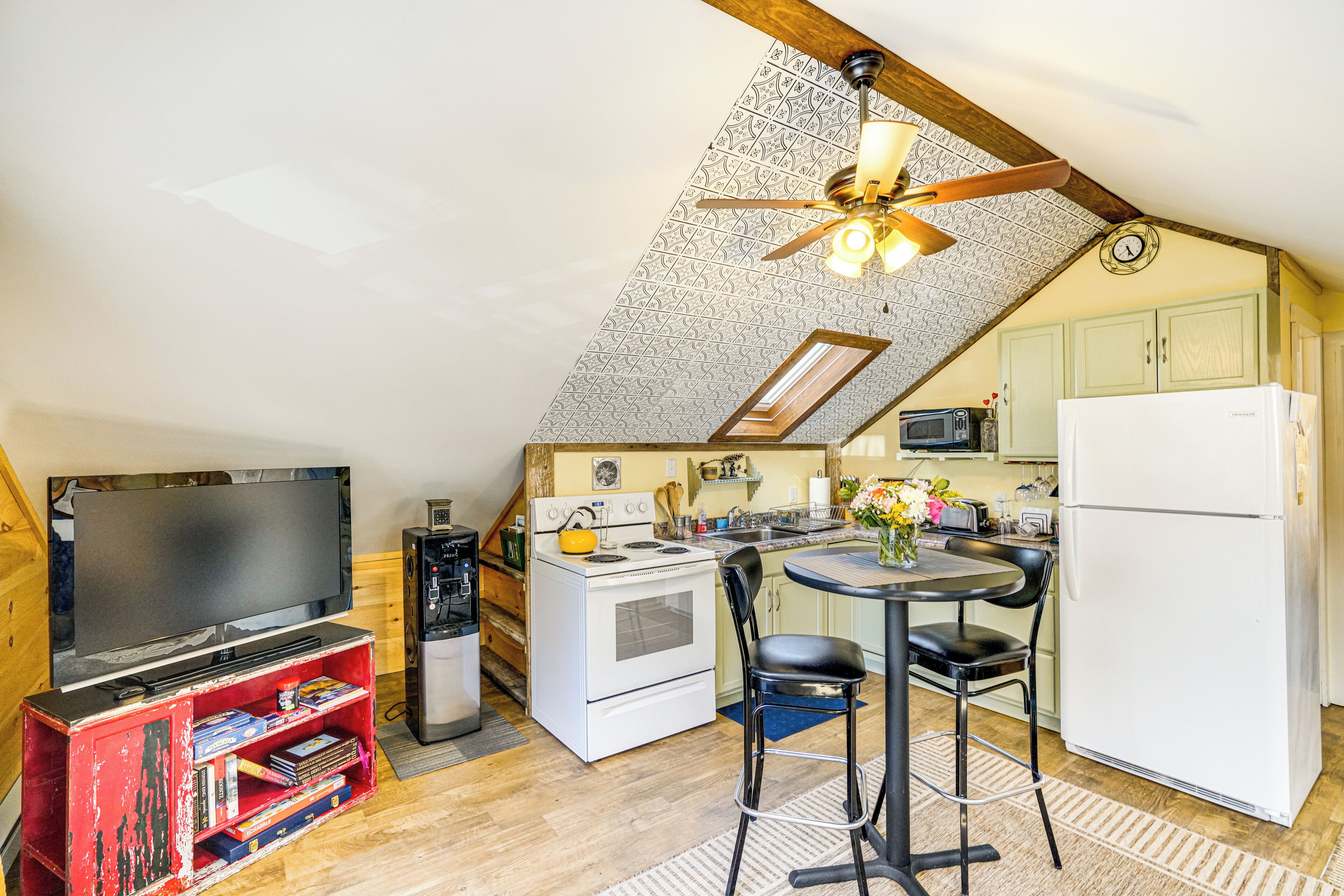 Une cuisine compacte équipée d’une cuisinière, d’un four à micro-ondes et d’un réfrigérateur. On y trouve une table à manger avec des chaises, ainsi qu’un téléviseur posé sur un meuble.