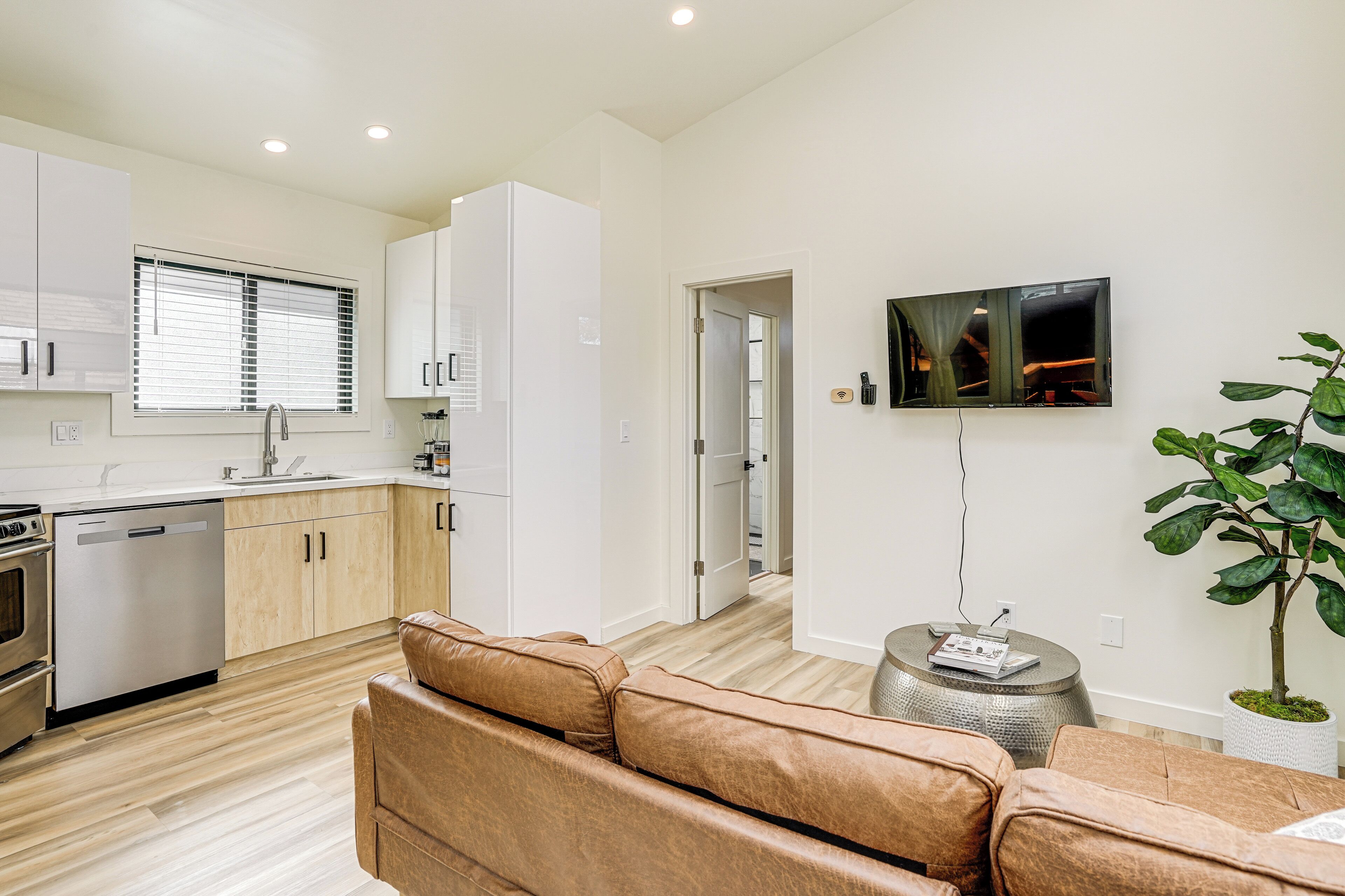 A modern kitchen with white cabinets, a built-in oven, and a dishwasher. There is a brown leather sofa in the living area, a round side table, and a potted plant.
