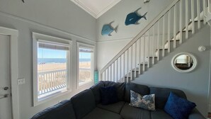 Ein Wohnzimmer mit einem grauen Ecksofa, zwei Fenstern mit Blick auf den Strand, einer Treppe mit weißen Geländern und zwei Hai-Wanddekorationen.
