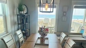 Ein Essbereich mit Holztisch, weißen Stühlen, einer Blumenvase und Blick auf den Strand durch ein großes Fenster.
