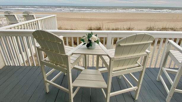Eine Terrasse mit zwei weißen Adirondack-Stühlen und einem kleinen Tisch, mit Blick auf einen Strand mit Wellen und Sand.