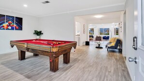 A pool table in a room with a wooden floor and white walls. There is a door on the right and a view of a living area with a sofa and a plant. - 6 Mi to Joshua Tree: Home w/ Outdoor Oasis & View! (Twentynine Palms)