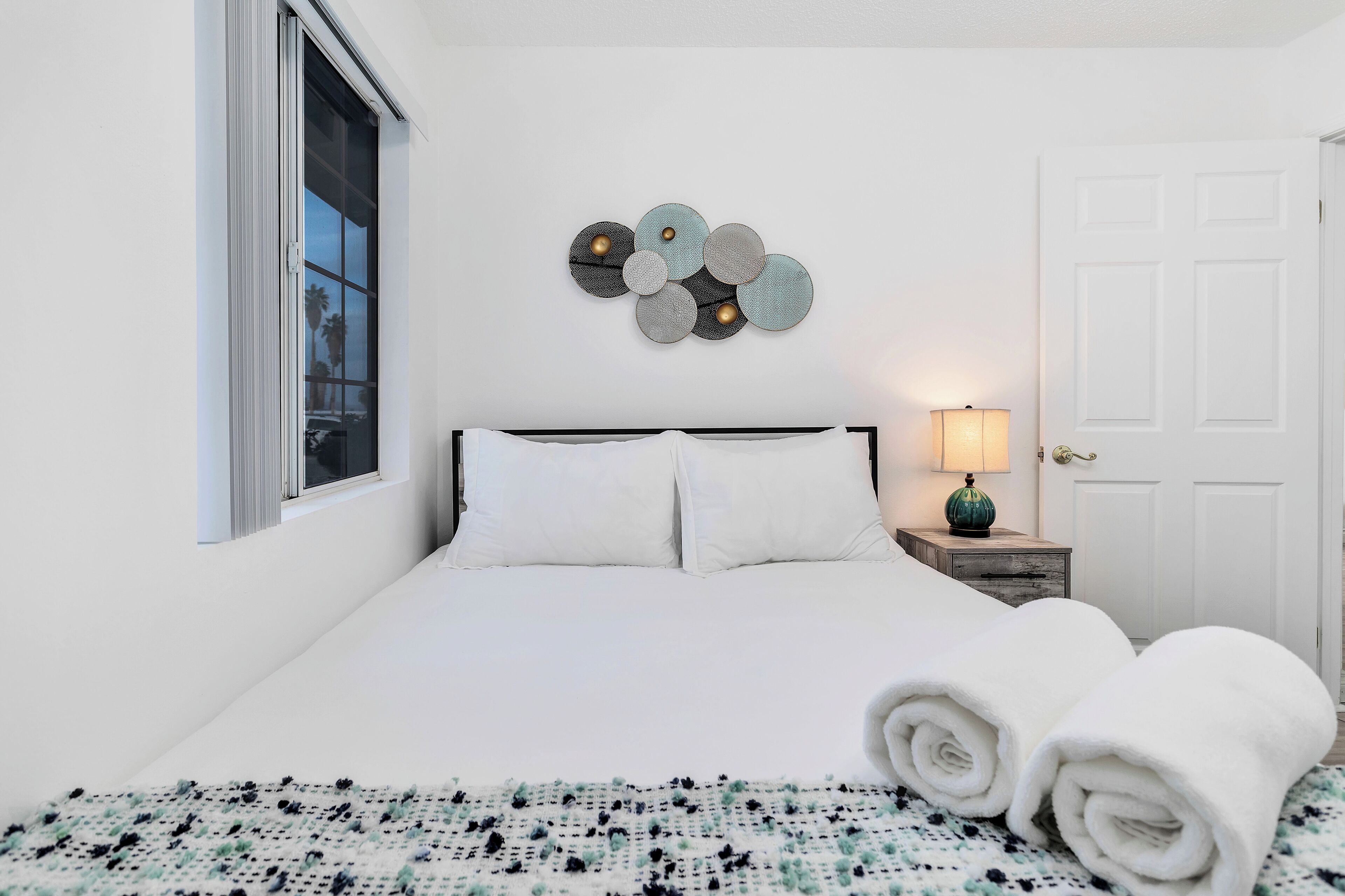 Een net opgemaakt bed met witte lakens, een decoratief kunstwerk erboven en een lamp op een nachtkastje.
