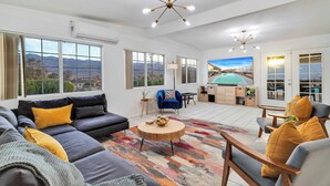 A spacious living room with a large sectional sofa, a blue armchair, and a wooden coffee table. There are multiple windows offering a view of the mountains. - 6 Mi to Joshua Tree: Home w/ Outdoor Oasis & View! (Twentynine Palms)