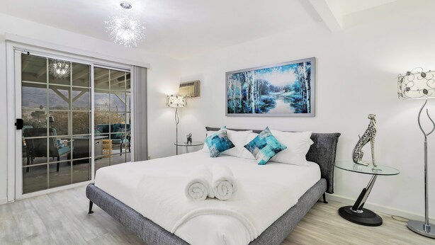 A modern bedroom with a large bed, a glass door leading to a balcony, a painting on the wall, and a small table with a decorative item. - 6 Mi to Joshua Tree: Home w/ Outdoor Oasis & View! (Twentynine Palms)