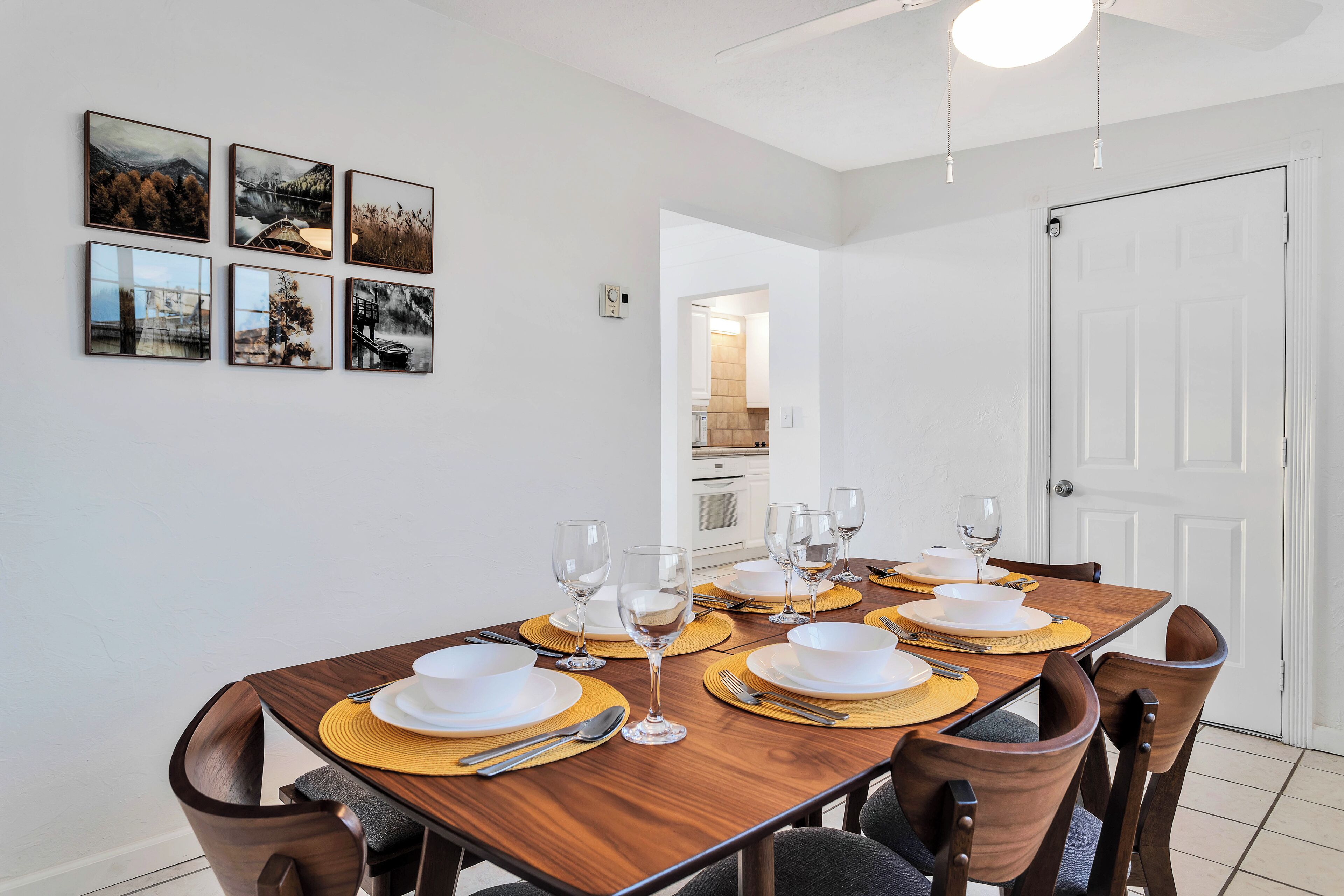 A dining area with a wooden table set for six, surrounded by chairs, with a kitchen visible in the background.