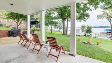 A covered patio with wooden chairs and a table, overlooking a grassy area and a body of water.