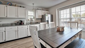 A modern kitchen with white cabinets, a dark wooden table, and a large window with blinds. - Charming 3 Bedroom Cottage in Bahama Beach Cottages! Dog Friendly, WIFI, Beach Town! (Bethany Beach)