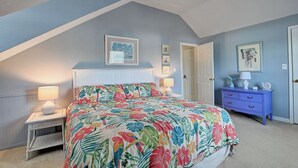 A bedroom with a sloped ceiling, a bed with a floral bedspread, a nightstand with a lamp, a dresser with decorative items, and framed pictures on the walls. - Charming 3 Bedroom Cottage in Bahama Beach Cottages! Dog Friendly, WIFI, Beach Town! (Bethany Beach)