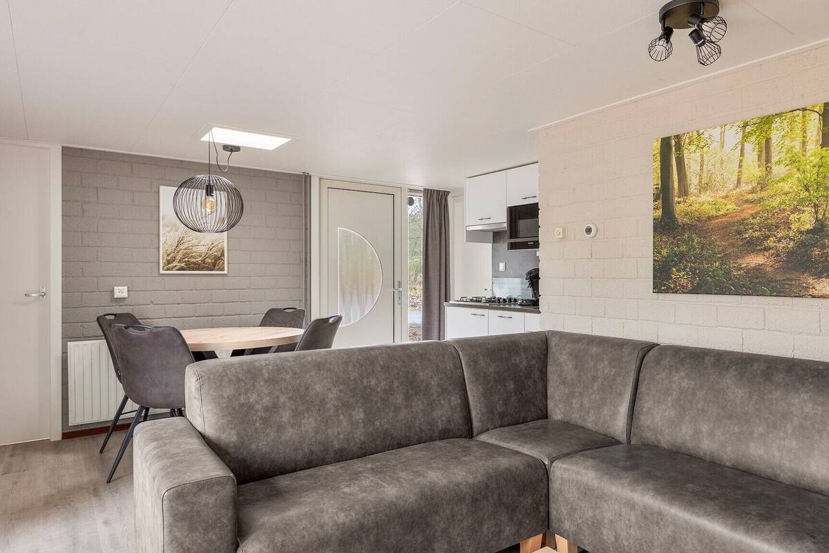 Un salon moderne avec un canapé d’angle gris, un coin repas avec une table et des chaises, une cuisine avec des meubles blancs et un grand tableau sur le thème de la forêt accroché au mur.