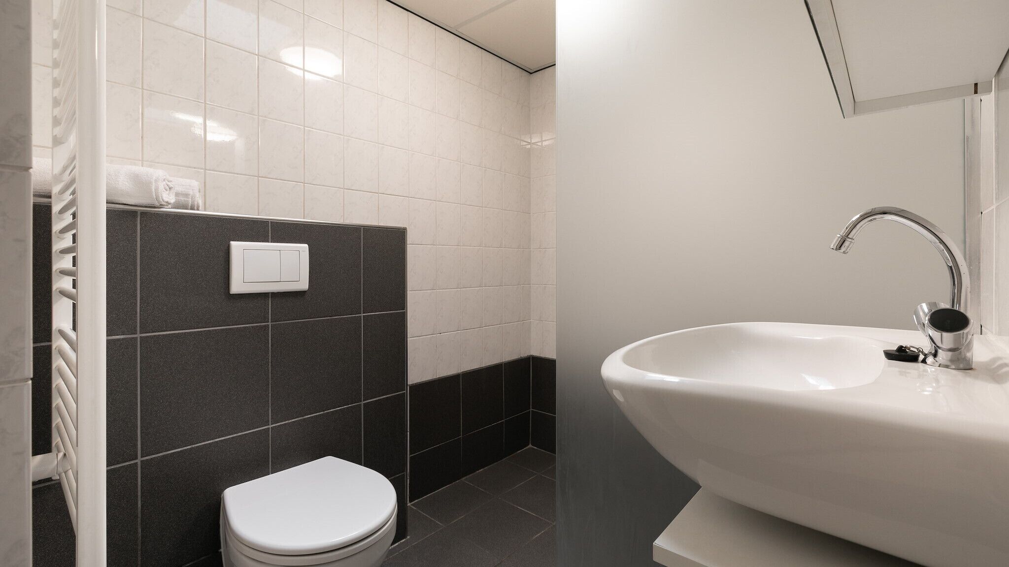 Een moderne badkamer met een wit toilet, een wastafel met chromen kraan en een lichtschakelaar aan de muur.