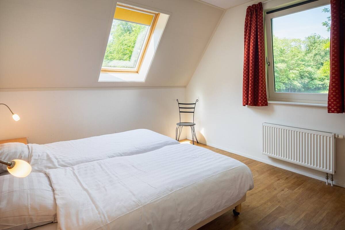 Une chambre à coucher avec un lit, une lampe de bureau, une chaise, un radiateur et une fenêtre avec des rideaux.