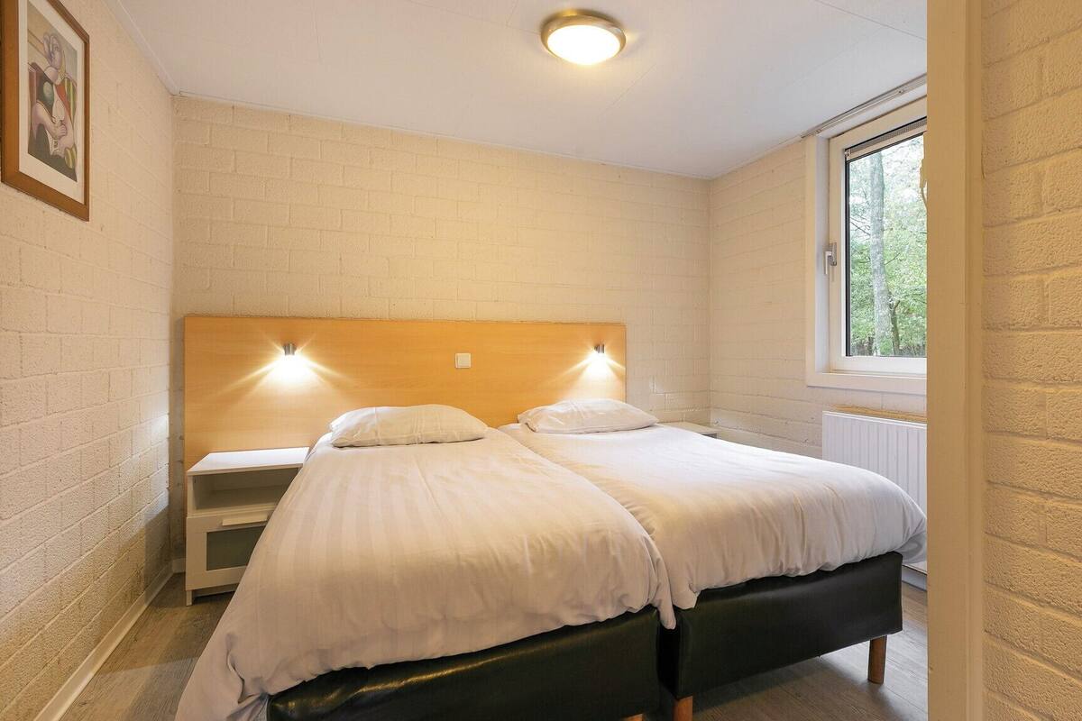 Une chambre avec deux lits, une tête de lit en bois, une fenêtre donnant sur des arbres et un mur en briques blanches.