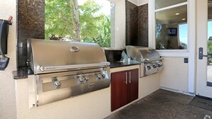 Cocina exterior con parrilla y estufa a gas, integrada en una vivienda con vista a árboles.
